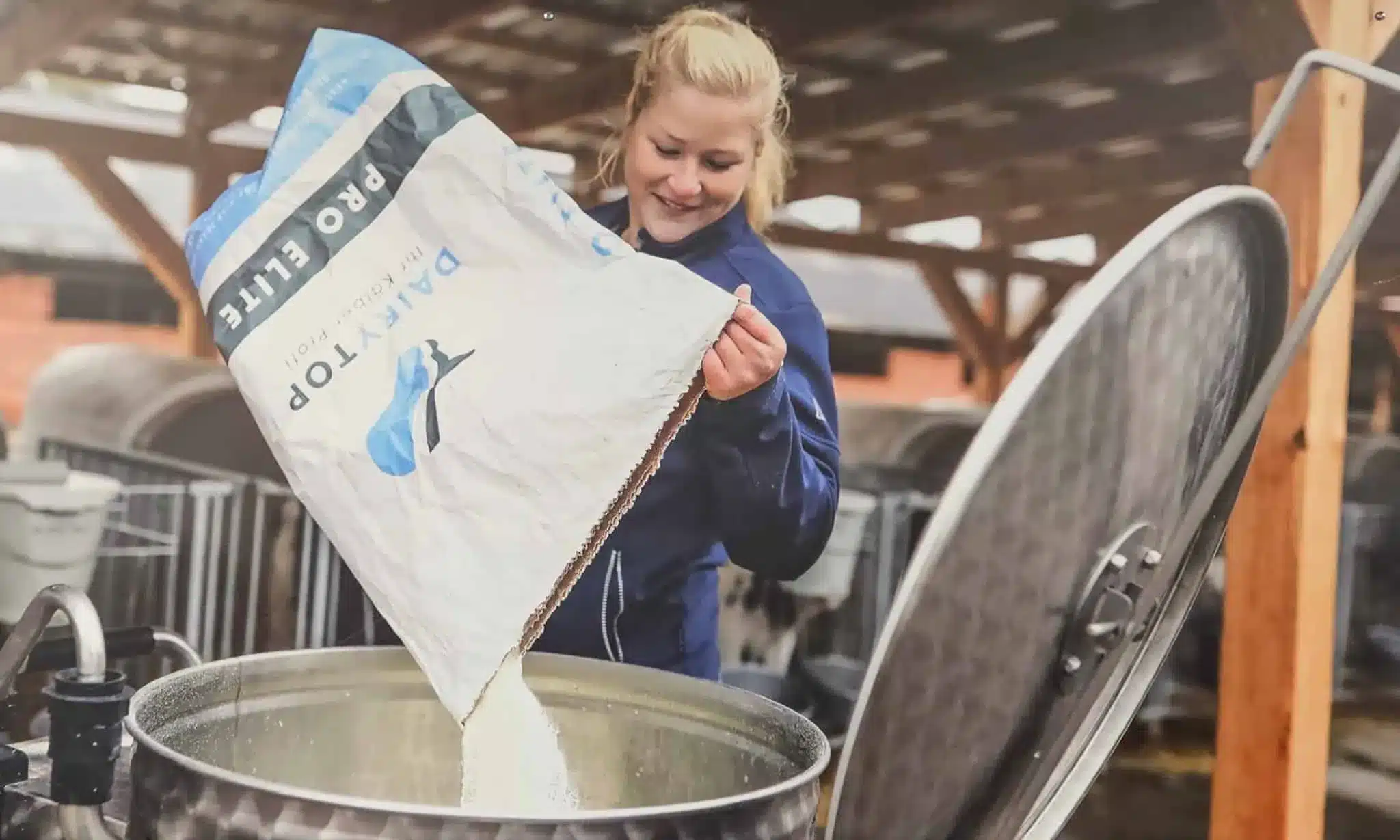 Een medewerker schudt een zak Pro Elite melkpoeder leeg in een melktank, met kalverhutten op de achtergrond